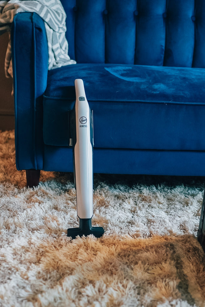 An upright vacuum cleaner with a gray and white color scheme and a black handle. The vacuum has a clear dustbin and a HEPA filter, and it is shown in use on a carpeted floor. There is a pet bed in the background with a gray and white patterned cushion.